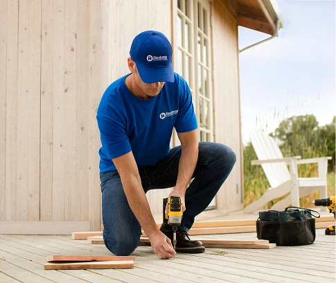 Technician in a blue Handyman Connection uniform kneels on a deck using a cordless drill, with boards and a tool bag nearby outside a wooden building.