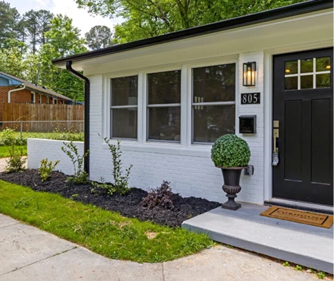 Freshly painted white brick house exterior with black front door, three windows, landscaped mulch beds, and the house number “805.”